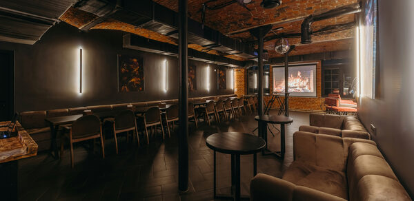 Авторские пространство T&S loft (ти энд си лофт) . Интерьер зала с кирпичными сводчатыми потолками и чёрными стенами. Длинные столы и стулья, отдельно стоящие круглые столики. Постеры и световые панели на стенах. Мягкие диваны, проектор с изображением камина. Теплое и приглушенное освещение.