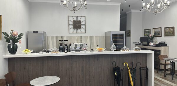 Арт Холл. Кафе с деревянной барной стойкой, круглые столы и стулья, светильники-подвески, светлые стены, картины, металлические зонтовницы с зонтами, кофемашина и напитки на стойке, декор в виде часов, деревянный пол.