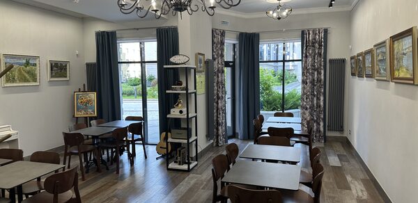 Арт Холл. Просторное помещение с паркетным полом и белыми стенами, украшенными картинами. Зал обставлен деревянными столами и стульями, освещен двумя люстрами. Вдоль одной стены расположены окна с синими шторами, межкомнатные перегородки отсутствуют.