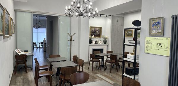 Арт Холл. Просторное помещение с деревянными полами, расставленными деревянными столами и стульями. На стенах картины. Две люстры с лампами. Стеклянные раздвижные двери ведут в другую комнату. Есть камин, полки с декором и стойка для одежды.