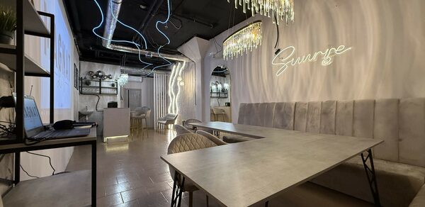 Лофт SUNRISE(Восход Солнца). Просторное помещение на 15 гостей. Прямоугольное помещение с длинным столом и стульями. Вдоль стены мягкие диваны. Потолочные светильники с хрустальными элементами. Светлая плитка на полу. На стене неоновая надпись. Открытые полки с растениями и техникой. Темный потолок с вентиляцией.