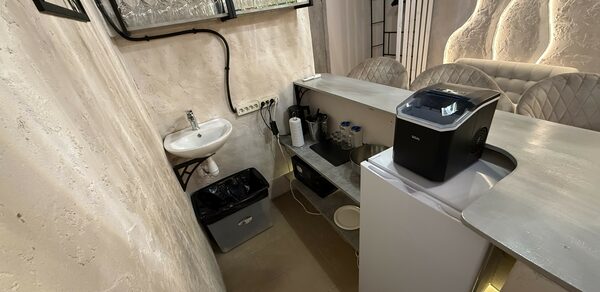 Лофт SUNRISE(Восход Солнца). Просторное помещение на 15 гостей. Маленькая кухня с барной стойкой, бежевые стены, белая раковина, металлические полки для бокалов, встроенный освежитель, мусорное ведро, белая техника, настенное освещение с изогнутыми линиями.