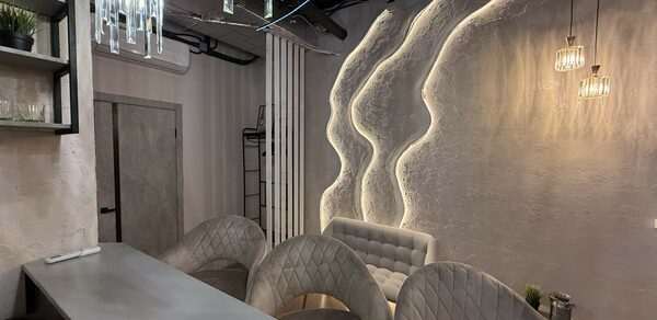 Лофт SUNRISE(Восход Солнца). Просторное помещение на 15 гостей. Современное помещение с барной стойкой и мягкими креслами, декоративное освещение с подвесными лампами, акцентная стена с текстурой и подсветкой, открытые полки с растениями, светло-серые и бежевые оттенки, минималистичный интерьер.