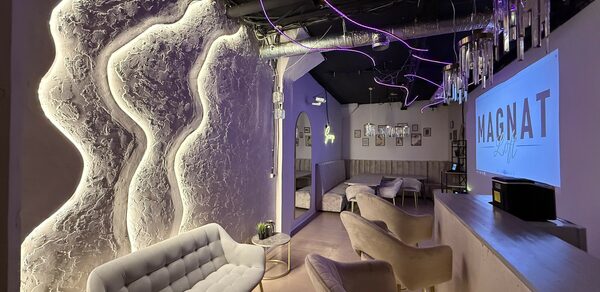 Лофт SUNRISE(Восход Солнца). Просторное помещение на 15 гостей. Лофт пространство с неоновым и белым освещением, выделяющим текстурированные стены. Мягкие стулья и диваны, столики. Барная стойка слева. Потолок с открытыми коммуникациями. На стене телевизор с надписью. Светильники свисают с потолка.