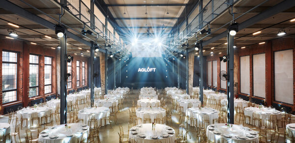 AG LOFT. Grand Hall (АГ Лофт. Большой зал) . Большое индустриальное пространство с высокими потолками и множеством круглых столов, покрытых белыми скатертями. Стулья прозрачные, на столах находятся свечи и бокалы. Огоньки сверху и кирпичные стены. На стене текст: "AGLOFT".