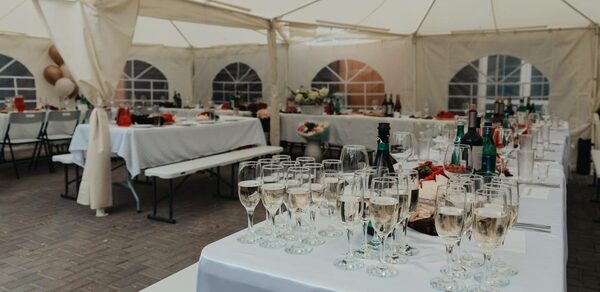 Усадьба Афонинская. Большой шатер с белым потолком и стенами, прозрачные окна полукруглой формы. Длинные столы, покрытые белыми скатертями, сервировка бокалами и бутылками. Ряды складных стульев и лавки. Пол из серых плиток. Декор из цветов и воздушных шаров.