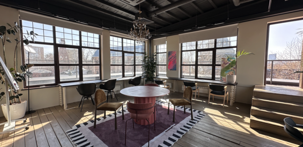 Пространство с террасой до 50 человек. Просторное помещение с большими окнами и деревянным полом. Центральный круглый стол окружен стульями на ковре с геометрическим узором. Подвесная люстра на черном потолке с видимыми трубами. Растения и картины по стенам.