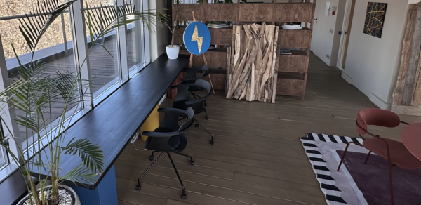 Пространство с террасой до 50 человек. Пространство в стиле лофт с большими окнами, вдоль которых стоит длинный стол с черными офисными стульями. На полу деревянный настил, коврик с черно-белыми полосами и красными элементами, разделение из деревянных панелей. Декоративные растения.