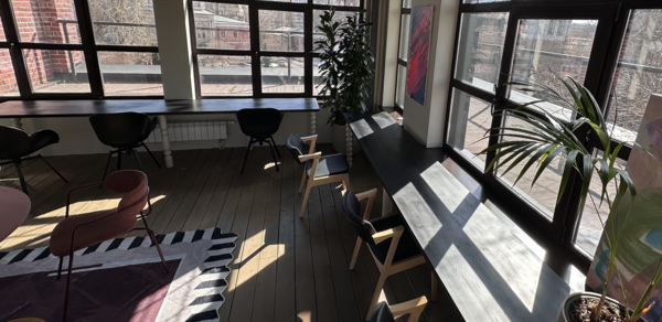 Пространство с террасой до 50 человек. Просторное помещение с большими окнами, светлое дерево на полу, черные и розовые кресла, длинные столы вдоль окон, несколько растений, абстрактная картина на стене, ковёр с геометрическим узором, кирпичная стена у окна.