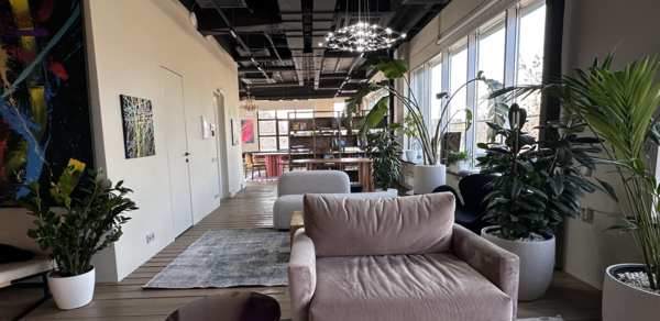 Пространство с террасой до 50 человек. Современное пространство для мероприятий с большим окном, серым диваном, фиолетовым столиком, множеством зелёных растений в кашпо. Потолок тёмный с видимыми вентиляционными системами, освещение в виде дизайнерских светильников, стены бежевые с художественными картинами.