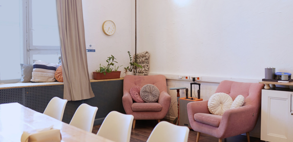 H4U. Светлый зал П-602. Современное пространство с панорамными окнами и кремовыми шторами. Обеденный стол из светлого дерева с белыми стульями. Два розовых кресла с подушками. Декор из растений и скульптур. Белые стены, лампа на стене, настенные часы.