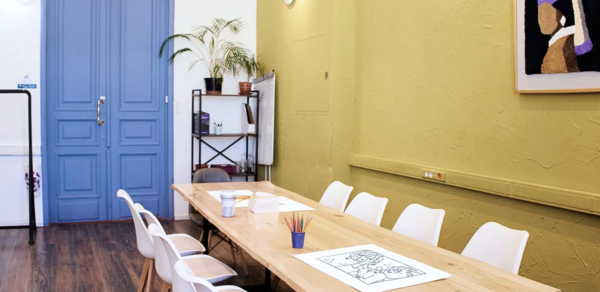 H4U. Светлый зал П-602. Пространство с длинным деревянным столом и белыми стульями. На стенах зелёная штукатурка, картина. Синяя двустворчатая дверь, тёмный деревянный пол. Полка с растениями и доска для записей. Верхнее и настенное освещение, цветные карандаши на столе.