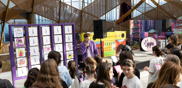 Кунцево Плаза. Современное пространство для мероприятий с высокими потолками и стеклянными стенами. Панель с рисунками и ведущей, выступающей у микрофона. Множество людей, в основном подростки. Цветная декоративная отделка и панели с изображениями.