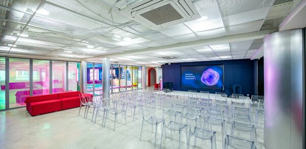 АКИ.лаб пространство для мероприятий. Современное концертное или презентационное пространство с прозрачными стульями, красными диванами, большим экраном на темной стене, бетонный пол, подвесной потолок с вентиляцией, разноцветная подсветка, стеклянные перегородки.