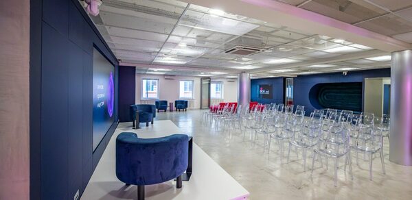 АКИ.лаб пространство для мероприятий. Просторное помещение с серым бетонным полом и подвесным потолком. Стулья из прозрачного пластика расположены перед подиумом с двумя синими креслами. Стены темные с большими экранами. Освещение встроенное, равномерное.