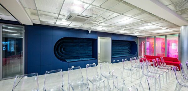 АКИ.лаб пространство для мероприятий. Просторное помещение с белым потолком и синей стеной, украшенной геометрическими панелями. Прозрачные пластиковые стулья расположены рядами. Справа стеклянная стена с розовой подсветкой. Пол светлого оттенка, потолок с встраиваемыми светильниками.