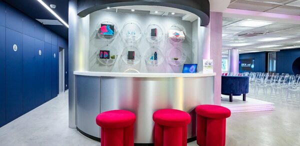 АКИ.лаб пространство для мероприятий. Современное помещение с белым столом полукруглой формы и тремя ярко-красными пуфами. Стены тёмно-синие с белыми дверями, пол серый. Подвесные светильники, стеклянные выставочные элементы на стене, прозрачные стулья, информационные таблички.