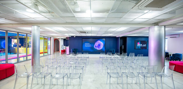 АКИ.лаб пространство для мероприятий. Просторное помещение с белым потолком и бетонным полом. Прозрачные пластиковые стулья расставлены рядами перед экраном на синей стене. Яркое электрическое освещение, стеклянные колонны, цветные стены сбоку, яркие текстильные диваны.
