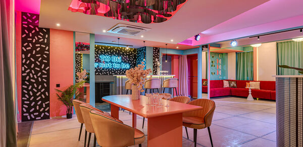 Лофт Шекспир. Стильный дизайнерский зал. Зал для мероприятий с ярким освещением, розовыми и бирюзовыми акцентами. Столы и стулья современных форм, барная стойка с высокими стульями. На стенах неоновая надпись. Декоративные панели с узорами, диваны с подушками. Потолок с декоративными элементами.