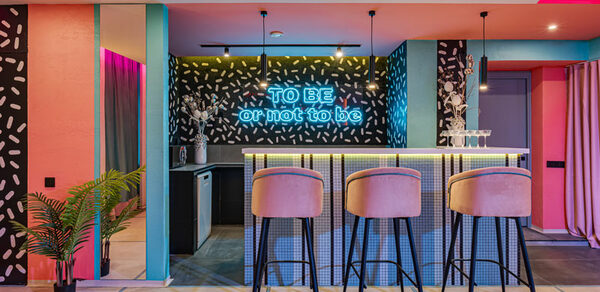 Лофт Шекспир. Стильный дизайнерский зал. Барная зона с высокими стульями и барной стойкой, подсвеченной неоновым текстом «TO BE or not to be». Стены с черно-белым узором, потолочное освещение, яркие цвета, элементы розового и голубого, зеркало, растения в углу.