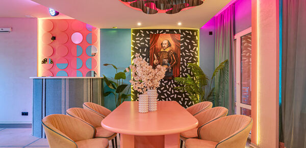 Лофт Шекспир. Стильный дизайнерский зал. Розовый овальный стол с бежевыми стульями, на стене портрет в рамке, барная стойка с круглыми элементами, розовый и голубой декор, растения, многоцветное освещение, шторы, потолочный элемент с волнениями.