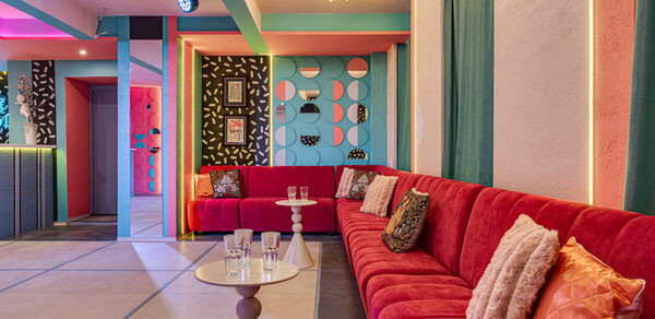 Лофт Шекспир. Стильный дизайнерский зал. Пространство с длинным красным диваном с подушками, круглый столик с бокалами, яркие стены с геометрическими узорами, различные картины, декоративное освещение, разноцветные стены и потолок, плиточный пол светлых оттенков.