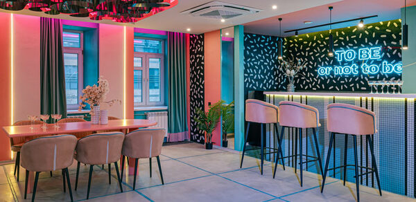 Лофт Шекспир. Стильный дизайнерский зал. Современное праздничное пространство с барной стойкой и столовой зоной. Бар с высокими стульями, неоновая надпись, обеденный стол с мягкими стульями. Яркое декоративное освещение, разноцветные стены, большие окна с шторами, растения в горшках.