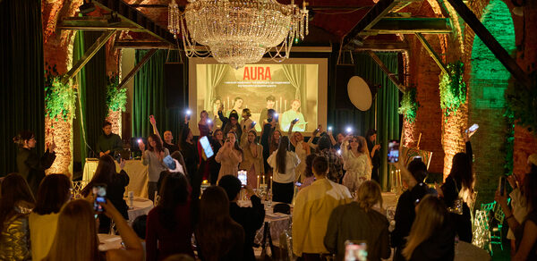 Arsenal Loft(Арсенал Лофт). Банкетный зал с кирпичными стенами и арочными потолками. Большая люстра, зеленые и оранжевые подсветки. Люди стоят у сцены. Круглые столы с белыми скатертями. На заднем плане экран с надписью "AURA".