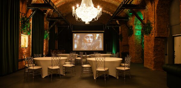 Arsenal Loft(Арсенал Лофт). Винтажное пространство с кирпичными сводчатыми потолками и стенами. Круглые столы с белыми скатертями, прозрачные стулья. Большая люстра и зелёная подсветка. Экран с проекцией. Тёмный пол. Живые растения на стенах.