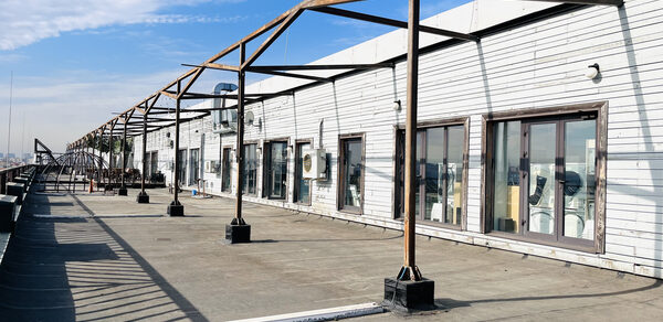 Зал под кулинарные мастер классы и мероприятия. Терраса под открытым небом с деревянным навесом, витражными окнами и кондиционером на белой стене здания. Деревянные балки поддерживают структуру, на полу бетон. Светлый день, частично облачное небо.