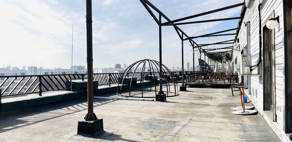 Зал под кулинарные мастер классы и мероприятия. Открытая терраса с бетонным полом, металлическими каркасами и перилами, деревянными столами и стульями справа. Видна часть здания с деревянной отделкой и кондиционером. Потолочные гирлянды на каркасах, дневное освещение.