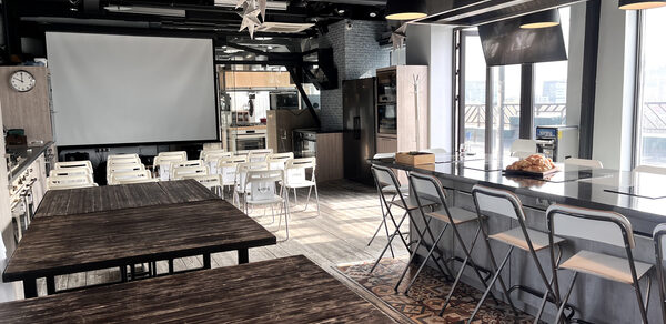 Зал под кулинарные мастер классы и мероприятия. Просторное помещение с деревянными столами и белыми стульями, большой экран на передней стене. Справа барная стойка с высокими стульями. Потолок с подвесными светильниками. На полу плитка с узором. Есть большие окна.