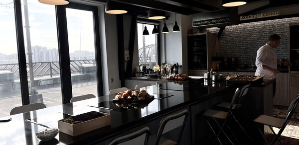 Зал под кулинарные мастер классы и мероприятия. Кухонное пространство с длинным островом из темного материала, окружённым складными стульями. На столе расположены блюда и столовые приборы. Подвесные светильники, большие окна с видом на город, светлые стены, зона приготовления с плитой и вытяжкой, текст на стене.
