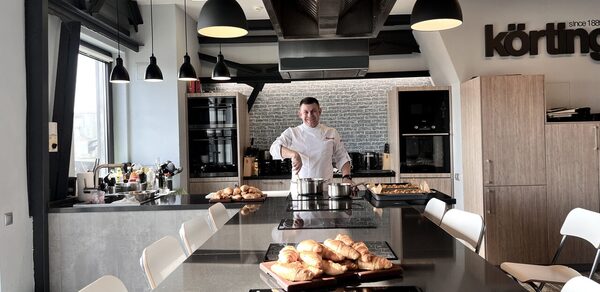 Зал под кулинарные мастер классы и мероприятия. Кухонное пространство с островом, варочная панель, современное освещение, подвесные светильники, барные стулья, деревянные шкафы, стеклянная дверь, выпечка на прилавке, человек в белой униформе, металлическая вытяжка, стена с кирпичным узором, логотип на стене.