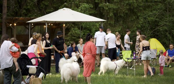 Белый Пёс(WHITE DOG event). Открытое пространство на лужайке, группа людей, белая палатка, стол и стулья, коляски, собаки. Зеленая трава, деревья на заднем плане. Яркие цвета одежды, желтая палатка слева.