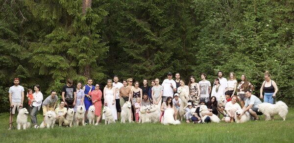 Белый Пёс(WHITE DOG event). Открытое пространство на фоне леса; множество людей и собак стоят на зелёной траве; высокие деревья с густой листвой; группа одета в летнюю одежду разнообразных оттенков.