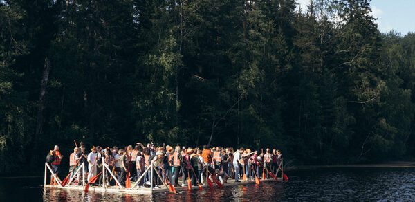 Белый Пёс(WHITE DOG event). Плот с множеством людей на спокойной воде озера, окружённого густым лесом. У людей спасательные жилеты и весла. Солнечный день, свет падает на плот, создавая игру теней на воде. Деревья густые, зелёные, сочные.