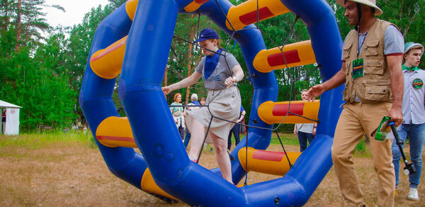 Белый Пёс(WHITE DOG event). Открытое пространство на лужайке с большим надувным кольцом синего и жёлтого цвета. В центре кольца человек в спортивной одежде. Вокруг стоят люди. На заднем плане деревья и небольшой белый домик.