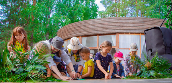 Белый Пёс(WHITE DOG event). Дети и взрослые сидят на деревянной сцене с зелеными растениями в руках. Овальное деревянное здание с большими окнами на заднем плане. На улице, вокруг зеленые деревья. Справа аудиоколонка и штатив с освещением.