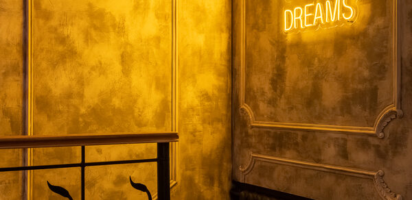 Каминный Зал Tangiers lounge Pokrovka(Танжерс Лаундж Покровка). Зона с декоративным металлическим ограждением и неоновой надписью на стене, текст читается частично. Стены с фактурной отделкой серого цвета, освещение теплое, желтоватое. Черный радиатор под стеной.