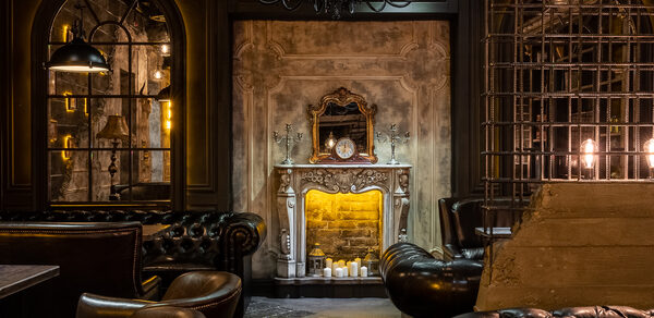 Каминный Зал Tangiers lounge Pokrovka(Танжерс Лаундж Покровка). Интерьерное пространство с темной атмосферой и кожаной мебелью. Центральный элемент — искусственный каменный камин с зеркалом, свечами и часами. Над камином — большой черный люстр. Показаны светильники с теплым освещением.