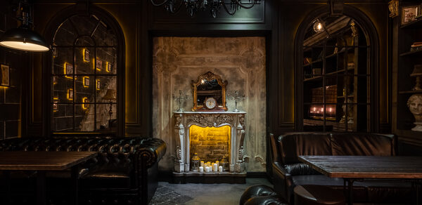 Каминный Зал Tangiers lounge Pokrovka(Танжерс Лаундж Покровка). Интерьер в классическом стиле с камином, кожаными диванами темного цвета и деревянными столами. Стены украшены книжными полками и статуей бюста. Потолочная люстра и несколько ламп накаливания создают теплое освещение.