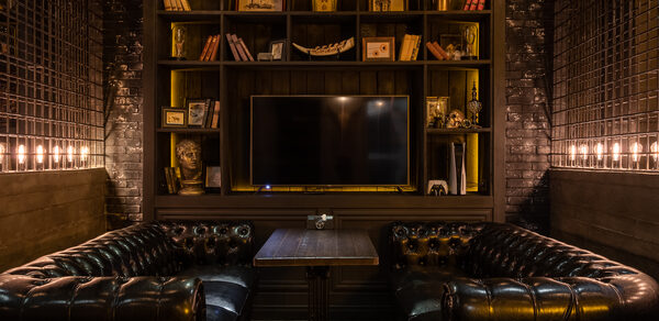 Каминный Зал Tangiers lounge Pokrovka(Танжерс Лаундж Покровка). Лофт-пространство с чёрными кожаными диванами, деревянным столом, стеллажом с книгами и декором, телевизором. Стены кирпичные, освещение тёплое, из лампочек над стеллажом и по бокам. Тёмная цветовая гамма, интерьер в классическом стиле.