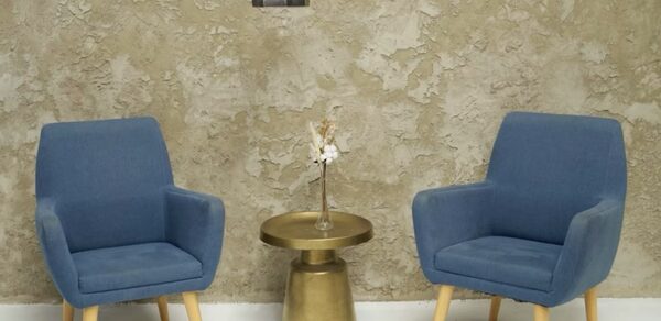 WhiteFox Studio(Студия Белая Лиса). Два синих кресла с деревянными ножками, маленький золотистый стол с вазой и сухими цветами между ними. Потолочный светильник над зоной, текстурная бежевая стена, белый пол.
