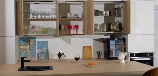 WhiteFox Studio(Студия Белая Лиса). Современная кухня с открытыми шкафами, наполненными посудой и бокалами. На столе бокал, тарелка с закуской, черный кран и раковина. Светлые стены, встроенная духовка, декоративные элементы и книги.