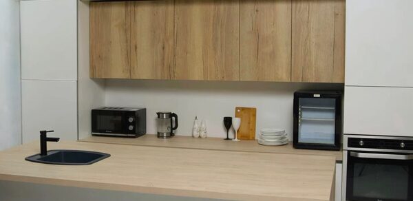 WhiteFox Studio(Студия Белая Лиса). Кухонная зона с деревянной столешницей и шкафами, встроенная техника: черная плита, микроволновая печь, мини-холодильник. Черная мойка и кран, электрический чайник, посуда, разделочная доска. Светлые стены и шкафы, контраст темной техники.