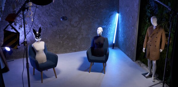 WhiteFox Studio(Студия Белая Лиса). Серое помещение с бетонными стенами, два манекена в креслах, один манекен в пальто стоит, стул с текстильной обивкой, неоновая лампа, потолочные и напольные светильники, фон из растений, светлая поверхность пола, студийное оборудование.