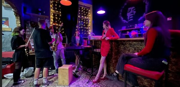 Лофт-Бар. Музыкальное пространство с барной стойкой, обставленное стульями и столиками, стены темные, на которых граффити. На стене висят световые гирлянды, освещение разноцветное. Пол выложен плиткой с узором.