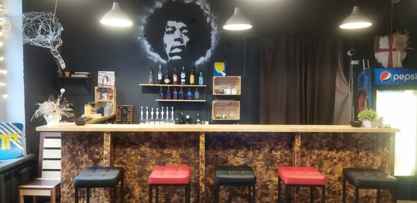 Лофт-Бар. Барная стойка с высокими стульями в черных и красных тонах, темная стена с изображением человека и полками с бутылками, подвесные светильники, холодильник с напитками, деревянные элементы декора, закрытая плотной темной шторой ниша.