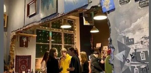 Лофт-Бар. Арт-пространство с обеденной зоной, стены украшены картинами, окно с гирляндами, столы и стулья из дерева, неоновые таблички, подвесные светильники, кирпичная кладка, разнообразные цветовые акценты, несколько человек общаются.